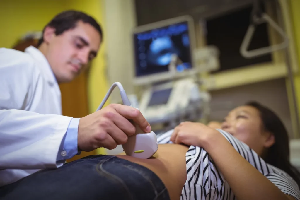 Médico realizando un ultrasonido a una mujer embarazada en un consultorio médico, con equipo de diagnóstico visible en el fondo.